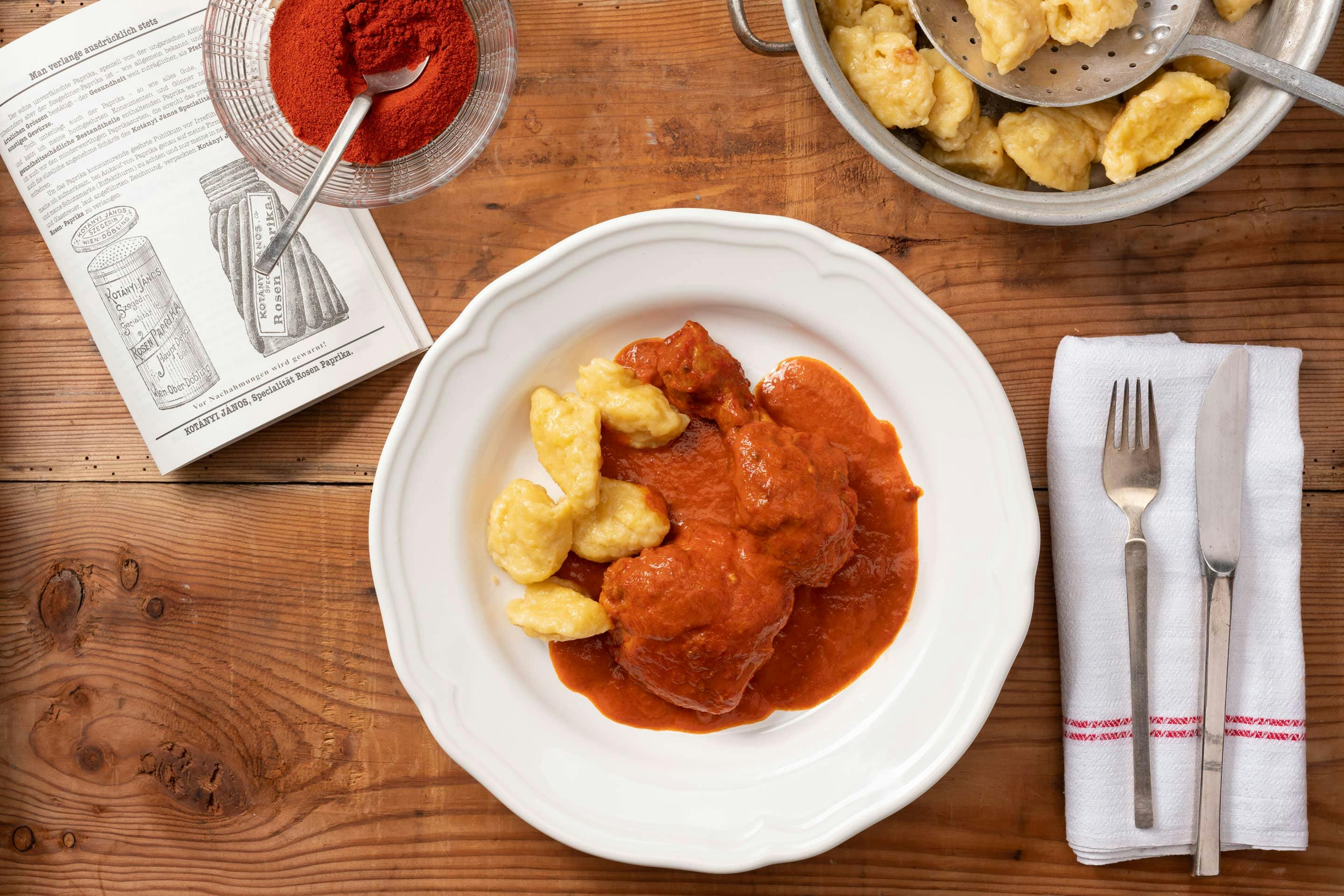 Глубокая тарелка с курицей с паприкой и клецками, рядом стоит миска с молотой паприкой.